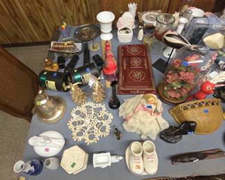 Table lot of collectibles including a glass display dome with faux hibiscus florals on a wood base, a carved shorebird on stand, a red lacquered Asian-style jewelry box with gilt medallions, Russian nesting-doll style figures, a milk glass urn vase, a vintage doll in lace gown, ceramic baby shoes, a cowrie shell mat, blue and white porcelain bud vases, and outdoor floodlight fixtures.