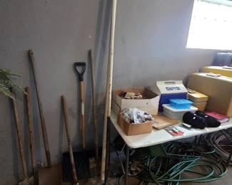 Garage wall of long-handled yard tools including shovels, a spade, snow shovel, and wood-handled implements, alongside a folding table holding an Igloo Playmate cooler, canning supplies, storage totes, and vintage hard-shell luggage, with coiled garden hoses below.