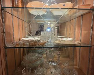 Lighted display cabinet with etched mirror-back doors holding peach luster ovenware, pink depression glass, cut and pressed glass bowls, crystal candlesticks, and hand-painted floral china serving plates. Multiple shelves of quality vintage glassware throughout.
