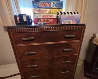 Art Deco waterfall-front chest of drawers with book-matched veneer, ribbed trim, and original hardware, topped with a stack of board games including Clue, Guesstures, Super Mario Monopoly, Scene It, Stratego, and DOS, plus a poker chip carousel and an automatic card shuffler.