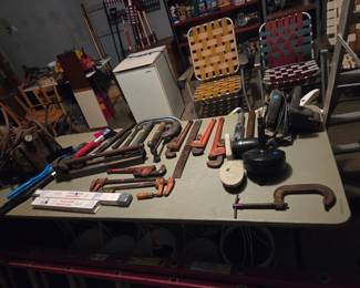 Workbench covered with hand tools and clamps, with folding lawn chairs and a small refrigerator nearby.