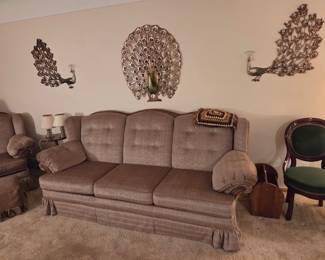 Beige tufted sofa with a skirted base, shown beneath a set of three decorative metal peacock wall sculptures.