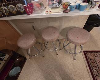 Set of three mid-century chrome swivel bar stools with round padded vinyl seats in a marbled taupe finish, shown at a laminate kitchen bar. Bar top behind holds Currier and Ives style wall plaques, coffee mugs, shot glasses, and a boxed serving caddy.