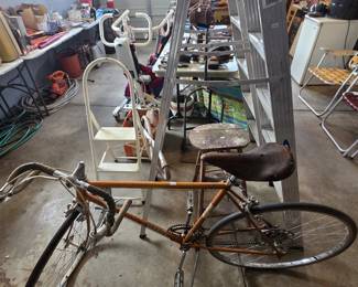 Vintage Varsity 10-speed road bike in orange with drop handlebars and a broken-in leather saddle, shown with a 6-foot aluminum stepladder, a white folding step stool, and a wooden milking-style stool.