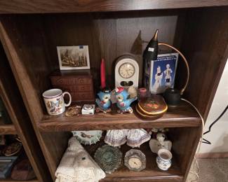 Bookshelf display of collectibles including a ceramic mantel clock, a miniature wooden jewelry chest with drawers, anthropomorphic bluebird figurines, a Delft-style porcelain boy and girl figurine set, a gooseneck desk lamp, crystal desk clock, crocheted lace tablecloth and doilies, a ceramic angel, and boxed lots of hand-painted Polish pisanki Easter eggs on the bottom shelf.