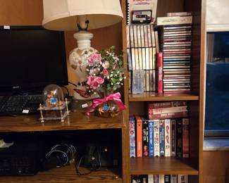 Closer view of the same bookshelf with books, VHS tapes, and a small ceramic table lamp.