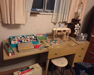 Vintage wood sewing machine cabinet with a matching stool, topped with sewing kits and craft supplies.