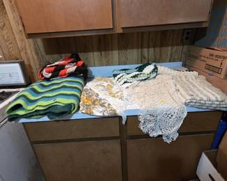 Counter of vintage textiles including a green and blue striped wool camp blanket, hand-crocheted lace tablecloths and doilies in ecru and white, a citrus-print round tablecloth with fringe, and several handmade afghans.