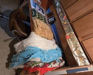 Table stacked with handmade crocheted afghans and fringed throws in white, aqua, red, and gold, plus Polish folk-print floral shawls with fringe; a rolled decorative metallic firescreen and a brown vinyl swivel chair nearby.