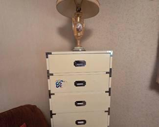 Cream campaign style six-drawer lingerie chest with brass corner brackets and recessed brass pulls, topped with an elegant gilt porcelain urn-form table lamp featuring a hand-painted courting scene and a fabric shade.