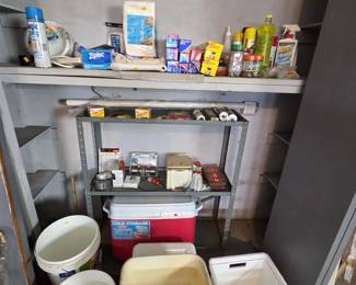 Garage pantry area with metal utility shelving and unopened household supplies including Ziploc freezer bags, Reynolds Wrap, Glad Cling Wrap, cleaning products, clothesline, paper plates, a Farmington Forge entrance lockset, a Rubbermaid 48-quart ice chest, and storage bins.