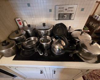 Large collection of vintage stainless steel cookware staged on the original General Electric built-in cooktop, including lidded stockpots, saucepans, skillets, a whistling tea kettle, and a black speckled enamelware roaster. Solid mid-century kitchen lot ready for daily use.