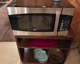 Small wood shelving stand holding a countertop microwave, with decorative dishware on the lower shelves.
