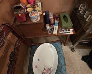 Two-tier mid-century telephone table topped with an assortment of pillar and jar candles, a decorative fruit-motif tin, an avocado green tissue box cover, beeswax candles, and new sewing notions. A vintage carved mahogany side chair in front holds a porcelain oval serving platter with pink rose decoration.
