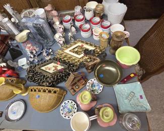 Tabletop of vintage decor featuring red-trimmed Santa mugs, hobnail milk glass vases, a cherub-decorated pitcher, ornate gold and black scrollwork frames with butterfly art, pink and green ceramic candleholders, wicker wall pockets, a carved crucifix, a brass bowl, souvenir plates, and a Swiss Army style pocket knife.