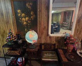 Large gilt-framed still life painting of yellow roses, an illuminated world globe on a carved gossip bench, a ceramic Christmas tree with lights, a black floral vase, a framed Last Supper print, a wall mirror, and a black metal rolling bar cart.