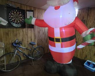 Large inflatable outdoor Santa Claus holding a candy cane, standing roughly eight feet tall and shown fully inflated and working. An electronic dartboard in a wall-mount cabinet and a vintage bicycle are visible in the background.
