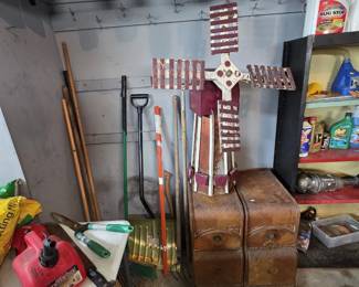 Handcrafted folk art wooden garden windmill in burgundy and white paint, displayed atop a pair of Art Deco waterfall-front cabinets with carved details and bracket feet; wall-mounted jars of sorted hardware overhead, long-handled garden tools, driveway markers, and a shelf of automotive fluids alongside.