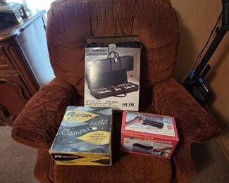 Upholstered rust chenille recliner holding a 40 inch keyboard carrying case model K-150 in box, a vintage Ricoh camera outfit box with leather case and flash unit, and a hand-operated paper shredder new in box.