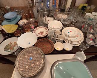 Dining table loaded with kitchen and entertaining pieces: a Farberware family-size griddle in the box, a Sunbeam electric skillet with turquoise lid, mid-century dinnerware with an atomic blue print, bud vases, stemware, wicker and copper baskets, milk glass serving bowls, and a woven sewing basket.