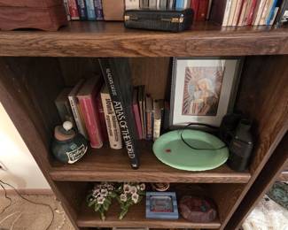 Bookcase shelves close up, featuring Polish-English dictionaries, a Readers Digest Atlas of the World, a framed Our Lady of Czestochowa style Madonna and Child print, a jadeite green glass oval platter, vintage binoculars, a Golfing Fund ceramic coin jar, and resin floral wall plaques.