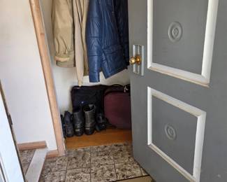 Entry closet with winter outerwear including a blue quilted parka, tan mens coats, and a knit sweater vest, plus insulated winter boots, a burgundy soft-side suitcase, and a black garment travel bag.