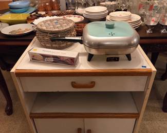 Vintage kitchen cart with a drawer and lower cabinet, topped with a mint-green electric skillet and colorful ceramic plates.