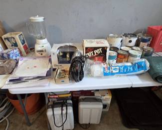 Long table of home improvement and kitchen items: a two-handle bathroom faucet new in box, West Bend Hot Pot heat-and-serve in original box, Black and Decker blender, vintage electric iron, Wilton paper doilies, box lot of small hardware, plastic sheeting, and an assortment of paint and stain cans; boxed Patton and Comfort Zone utility heaters underneath.
