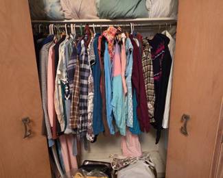 Closet filled with vintage womens clothing on hangers, including plaid flannel shirts, sweaters, blouses, and jackets, plus a shelf of blankets, quilts, and bed linens and boxes of handbags and knitwear below.