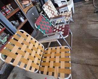 Collection of vintage aluminum folding lawn chairs with woven webbing in harvest gold, brown and white, red and green, and burgundy colorways, plus white resin stacking patio chairs; garage workbench with Craftsman toolboxes, a metal file cabinet, and wooden tennis rackets in the background.