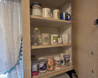 Corner cabinet of collectible mugs and glassware including a Grandma mug, a Cadillac Conner Stamping mug, a Life is Good mug, a golf mug with club handle, a playing card mug, a Coca-Cola glass, patriotic USA glasses, a fruit print glass, and a Branson Missouri souvenir mug.