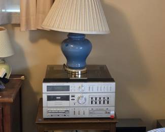 Sears AM FM stereo music system with turntable under a smoked dust cover, cassette recorder, graphic equalizer sliders, and auto-reverse tape deck, topped with a slate blue ceramic ginger jar table lamp with pleated shade, all sitting on a turned-leg wooden side table.