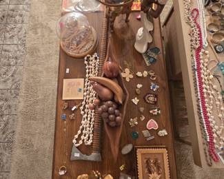Coffee table styled with mid-century carved wood pieces including a teak leaf-form centerpiece bowl with carved wooden fruit and grapes, a carved wooden horse, gilded clasped-hands sculpture, cowrie shell necklaces, a glass cloche dome, a carved Polish folk art wooden box with edelweiss motif, and an assortment of brooches, pins, and embroidered patches.