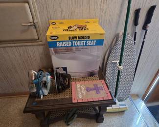 New in box Carex blow molded raised toilet seat, two vintage electric irons, a dark wood side table with woven cane inset top, a fabric-covered ironing board, sponge mop, ski poles, and an Easter Blessings burlap wall hanging.