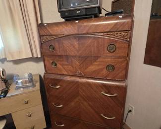 Art Deco waterfall chest of drawers from the 1930s-40s with bookmatched veneer front, carved scrollwork trim, round brass ring pulls, and shaped feet; a Zenith color television with remote sits on top.