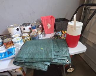 Painting and outdoor supplies including cans of Behr and Glidden paint, Minwax Gel Stain, Krylon rust preventive spray, folded green poly tarps, a pink daisy wastebasket, embossed metal planter box, red and white Coleman-style beverage jug, garden pinwheel spinners, a two-wheel hand truck, and rubber auto floor mats.
