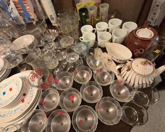 Large selection of vintage stemware and china including a set of etched glass champagne coupes and sherbets, milk glass mugs, colorful plastic tumblers, gold-rimmed cordial glasses, a brown drip-glaze teapot, a white and gold striped teapot, a Pepsi-Cola glass, and mid-century dinner plates with blue atomic pattern.
