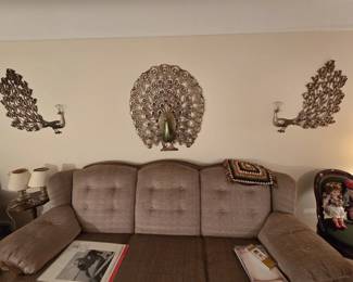 Striking three-piece vintage 1970s metal peacock wall art set, featuring a large full-fan center peacock flanked by two side-profile peacocks with jeweled tail accents. Displayed above a button-tufted sofa with a granny-square crochet pillow.
