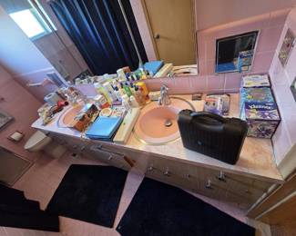 Retro pink tile bathroom with double-sink vanity holding grooming and personal care items including a Homedics case, hair dryer, curling irons, a Walgreens instant ear thermometer new in package, toiletries, and multiple boxes of facial tissue.