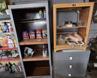 Gray metal utility cabinet with shelves holding WD-40, jars of sorted nuts and bolts, and a vintage circular saw, next to an Art Deco wood cabinet shell filled with paint rollers, brushes, and hardware, sitting on a gray three-drawer shop chest.