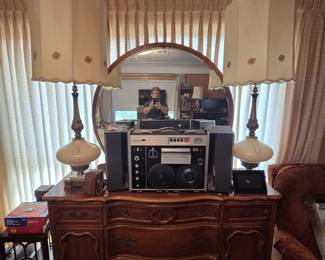 French Provincial serpentine-front triple dresser in cherry finish with scalloped door panels and brass pulls, topped by a round beveled wall mirror in a wood frame. A Sony reel-to-reel tape recorder with fold-out speakers sits on top, flanked by a pair of tall mid-century table lamps with glass bodies and original shades.