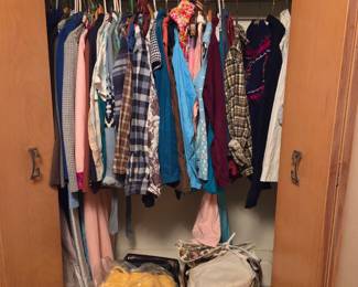 Closet full of hanging clothing with folded bedding on the shelf above and storage boxes on the floor below.