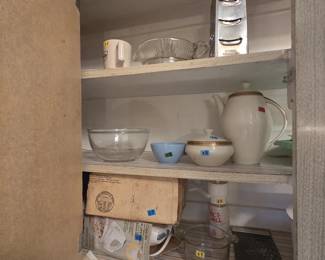 Upper cabinet with a porcelain coffee pot with gold band trim and matching lidded sugar bowl, a clear glass mixing bowl, a light blue bowl, a boxed vintage Sunbeam Mixmaster, a Nut-Mill grinder, a stainless box grater, and a glass measuring cup.