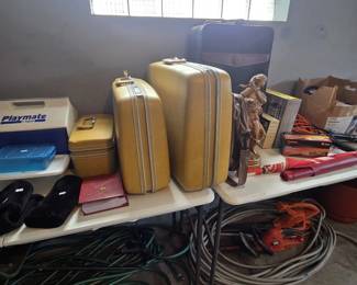 Vintage harvest gold hard-shell luggage set with two suitcases and a matching train case, plus a navy soft-side suitcase, Igloo Playmate cooler, bronze-finish figurine of a woman, AT&T Trimline 210 phone in box, and red photo albums; Black and Decker electric hedge trimmer and blower with garden hoses beneath the table.