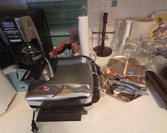 Vintage chrome waffle iron and sandwich grill with flip griddle plates, shown with a copper two-tier tidbit serving tray with brass center handle, aluminum garlic presses, a wood mug tree, milk glass bud vases, and clear glass tumblers.