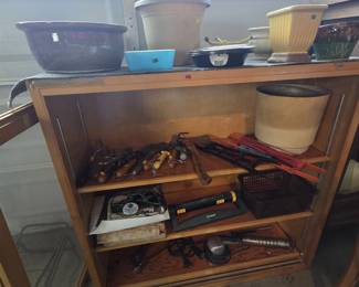 Wood bookcase topped with planters including a yellow ceramic footed planter, a large glazed bowl, and a turquoise blue refrigerator dish; shelves hold garden hand tools, a hatchet, wood-handled trowels and cultivators, a Melnor oscillating sprinkler, soldering supplies, and an electric engraving tool.