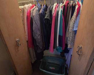 Closet filled with ladies clothing including wool coats, blazers, jackets, blouses, skirts, and slacks in a range of sizes and colors, plus a plastic laundry basket.
