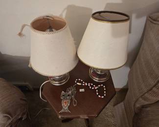 Hexagonal-top mahogany lamp table displaying two glass-font boudoir lamps with shades, a large pewter-tone articulated owl pendant necklace, and a red, white, and blue beaded necklace.