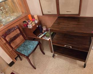 Mid-century rolling bar cart with a drop-front cabinet, shown beside a carved wood side chair with teal upholstery.