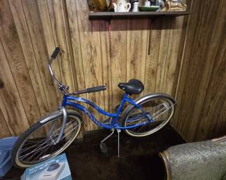 Vintage ladies cruiser bicycle in blue with fenders, whitewall-style tires, upright chrome handlebars, and a padded saddle. A shelf of collectibles above includes carved wood figures, ceramic figurines, a gold-tone dog statue, and a Kaz cool moisture humidifier in the original box.